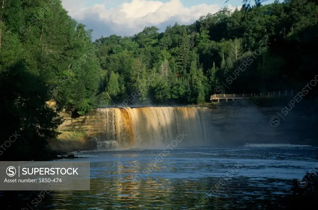 Usa, Michigan , Chippewa, Tahquamenon Falls Waterfall Surrounded By Lush Greenery And Man Standing On Riverbank Nearby