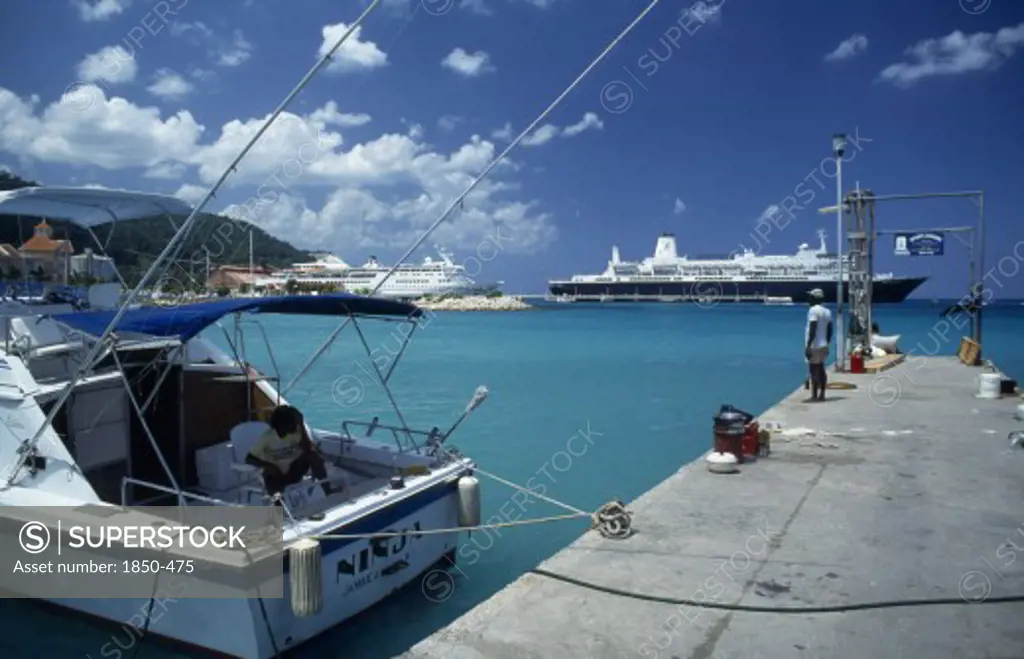 West Indies, Jamaica, Ocho Rios, Harbour View With Man Standing On Stone Jetty And Cruise Ships Behind.  Moored Boat In The Foreground With Person Sitting In Shade On Deck.