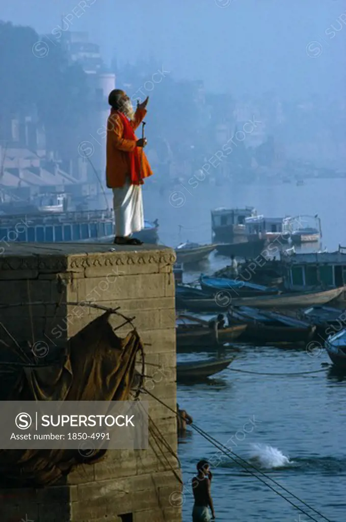 India, Uttar Pradesh, Varanasi, 'Holy Man Praying On Stone Wall Above River Ganges,Boats,Town,Mist '