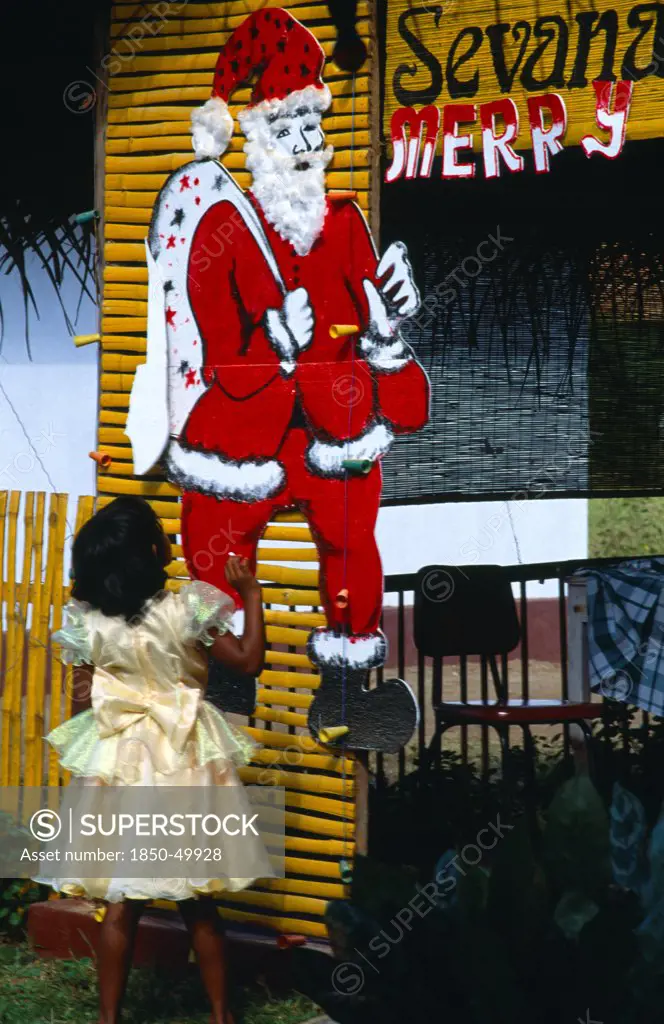 Sri Lanka, Religion, Christianity, Little girl in yellow party dress looking up at large cut-out of Father Christmas pinned to yellow painted frame.