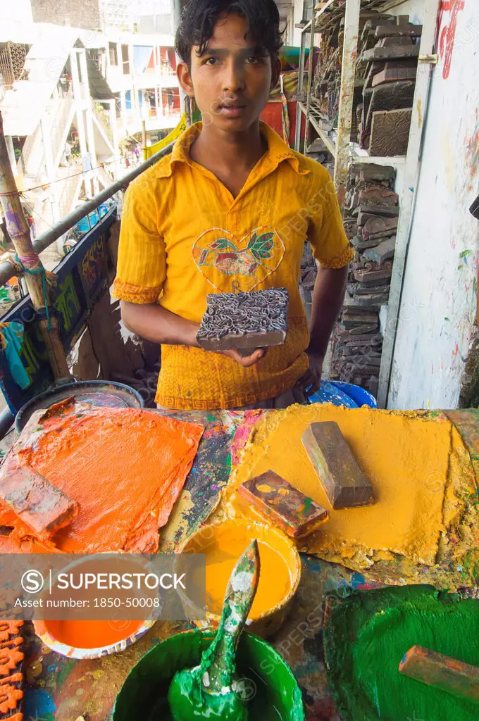 Bangladesh, Dhaka, Young man holding a printing block used to print on cotton fabrics in New Market with pallettes of colours in front of him.