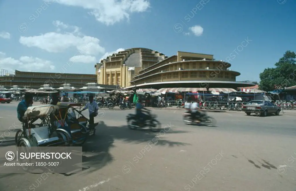 Cambodia, Phnom Penh, Trishaws And Motorcyclists On Road In Front Of The Central Market Buildings.