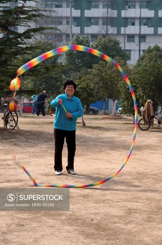 China, Jiangsu, Nanjing, Woman in a blue sweater and sneekers relaxing in a municipal park spinning a top with a multicoloured tail with an apartment block in the background.