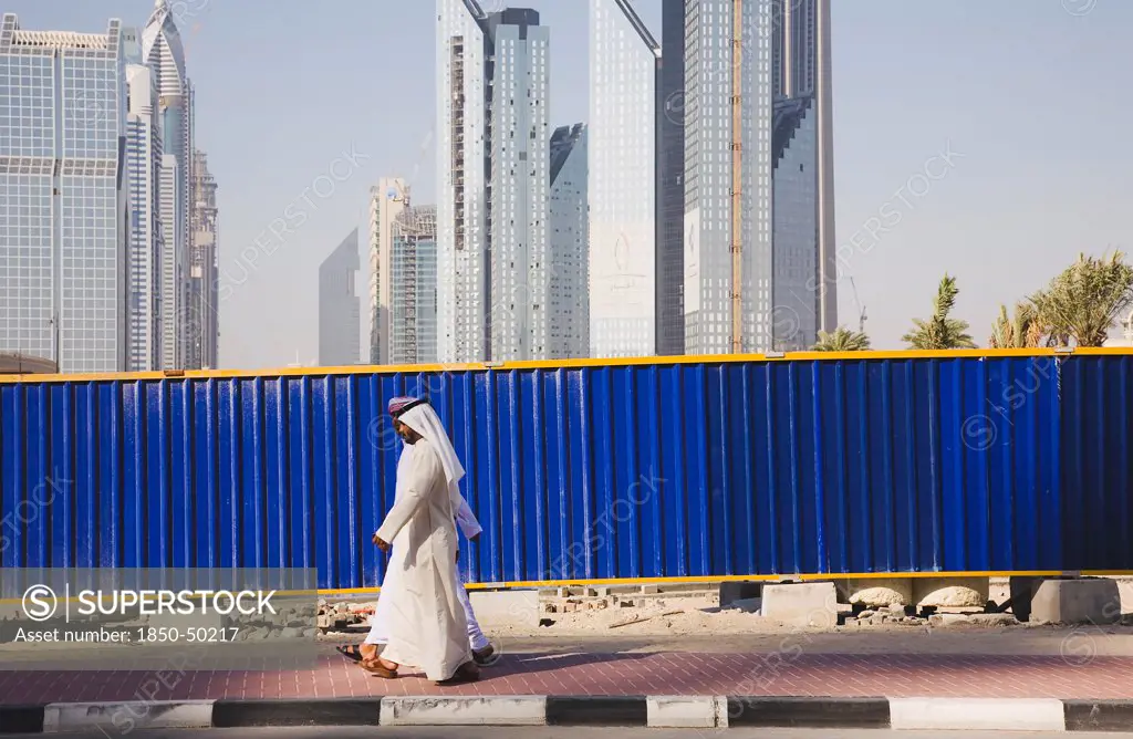 UAE, Dubai, Arab men dressed in tradityional Dishdasha walking past construction site with Sheikh Zayed Road behind.