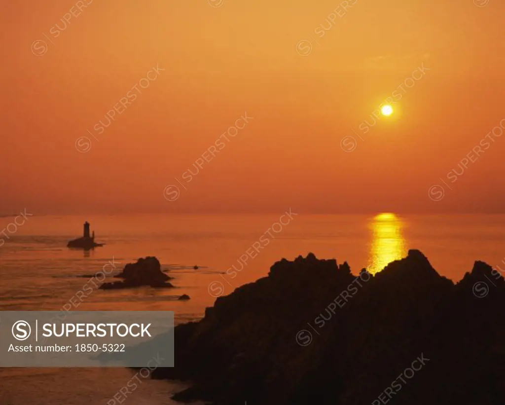 France, Brittany, Finistere, Near Audierne.  Pointe Du Raz At Sunset.  Rocks Silhouetted Against An Orange Sun And Evening Sky Reflected In The Water.