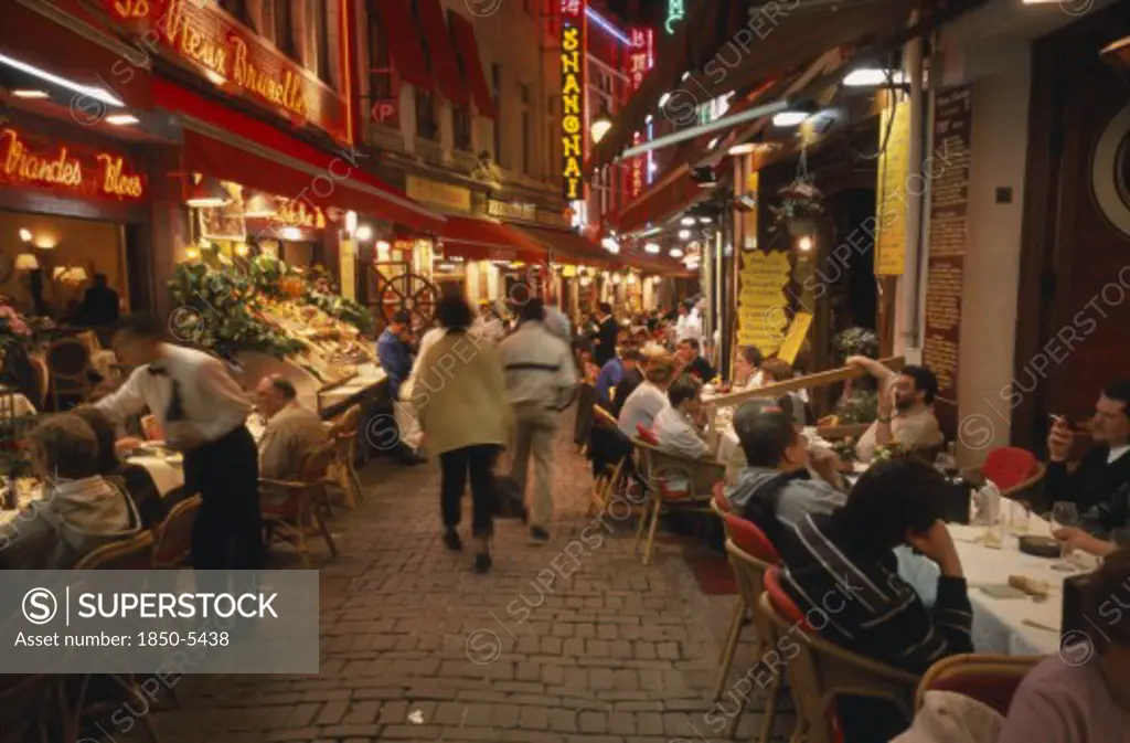 Belgium, Brabant, Brussels, People Sitting At Outside Tables In Narrow Street Lined With Restaurants At Night.
