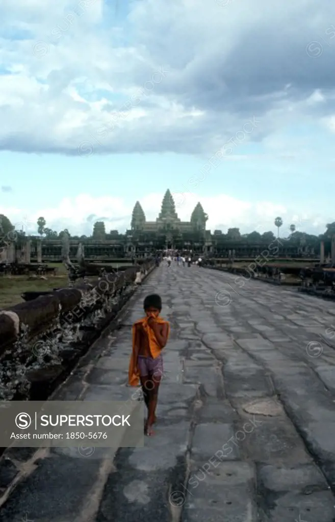 Cambodia, Siem Reap, Angkor Wat, Small Boy On The Stone Path Leading To The Temples Which Stand Against The Sky Behind.