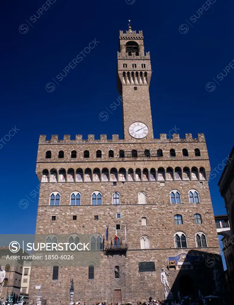Italy, Tuscany, Florence, Palazzo Vecchio.  Facade Of Medieval Palace Completed In 1322 And Used As The Town Hall.  Crenellated Roof And Clock Tower With Statues Outside Entrance Including Copy Of Michelangelos David.
