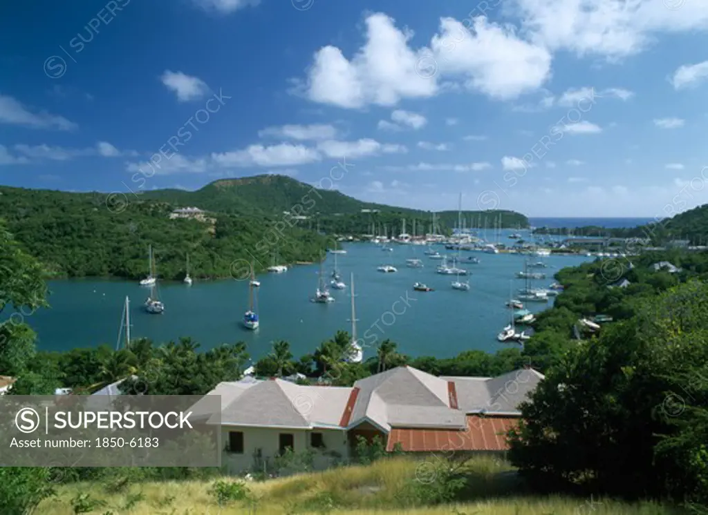 West Indies, Antigua, English Harbour, Ordnance Bay.  View Over Rooftops Towards Bay With Moored Yachts And Surrounding Tree Covered Coastline.