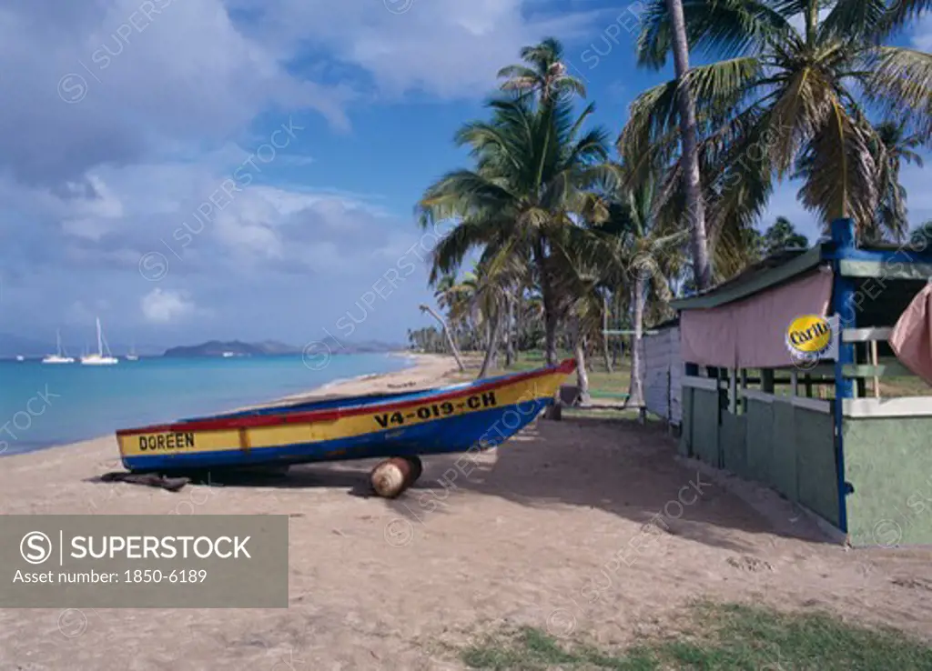 West Indies, Nevis, Pinneys Beach, Empty Sandy Beach Fringed By Palm Trees With Painted Wooden Boat Pulled Up Out Of The Water Beside Empty Hut Advertising Carib Beer.