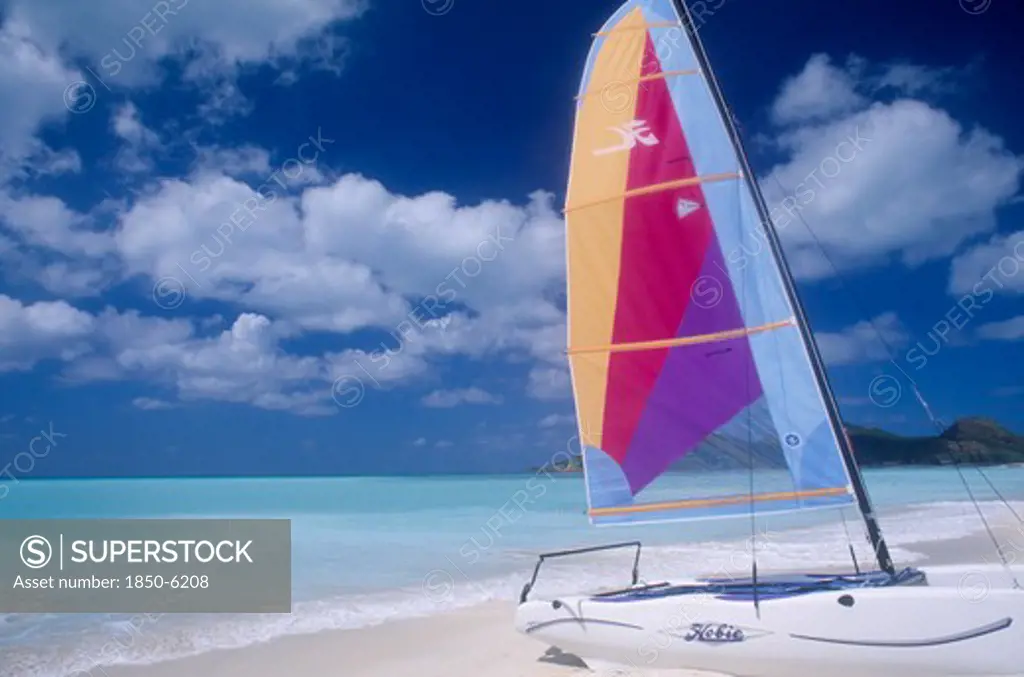 West Indies, Antigua, Jolly Beach, Empty Sandy Beach With Hobiecat With Brightly Coloured Sail In The Foreground.  Flat Calm Aquamarine Sea And Cloudscape Behind.