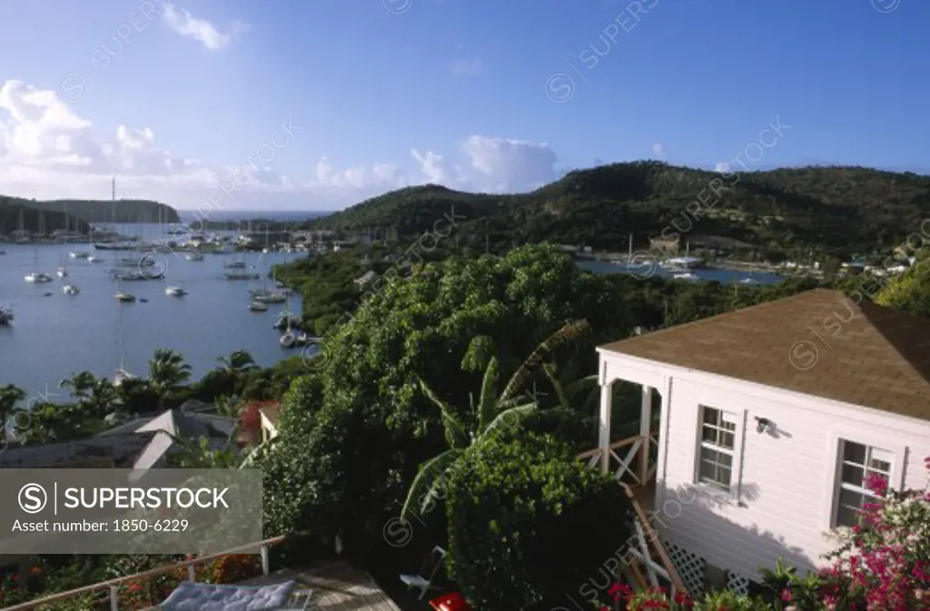 West Indies, Antigua, English Harbour, Ordnance Bay.  View Over Bay With Moored Yachts And Tree Covered Coastline With Part Seen White Painted House In The Foreground.
