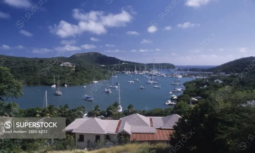 West Indies, Antigua, Ordnance Bay, View Over House Rooftops Towards Bay With Moored Yachts And Surrounding Coastline.