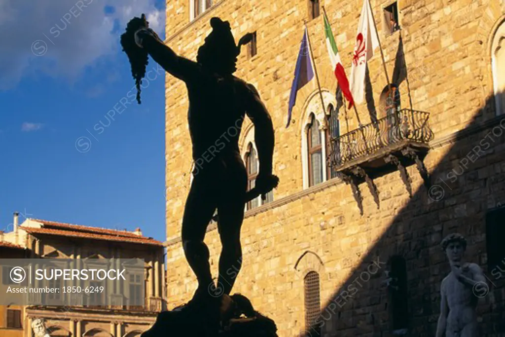 Italy, Tuscany, Florence, Piazza Della Signoria.  Statue Of Perseus Holding The Head Of Medusa By Cellini Silhouetted Against Palazzo Vecchio In Sunlight.
