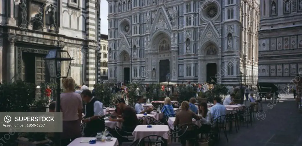 Italy, Tuscany, Florence, Cafe In Front Of The Neo-Gothic Marble Facade Of The Duomo With People Sitting At Outside Tables Having Coffee And Reading Guide Books.