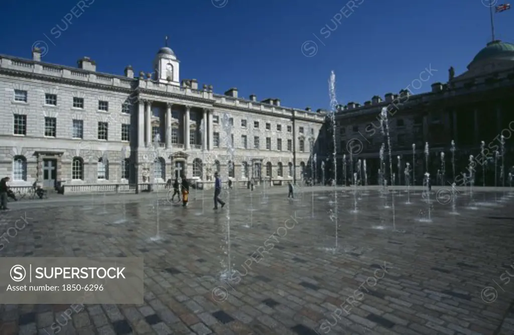 England, London, Somerset House And Courtyard With Rows Of Fountains Spouting High From The Ground With People Walking Between Them. Flag Flying At Half Mast In The Distance.
