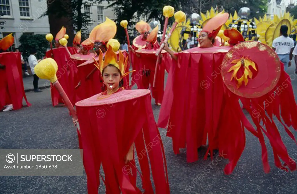 England, London, 'Notting Hill  Carnival. Little Girl In Red Costume And Flame Shaped Head-Dress, Adults In Costume Blowing Whistles Behind Her. '