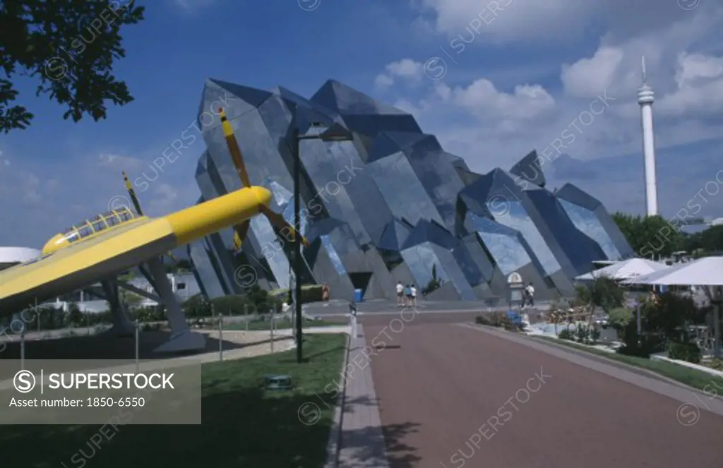 France, Poitiers, Planet Futuroscope, Le Parc Europeen De LImage. Sur Les Traces Du Pamda. Path Leading To Mirrored Modern Architecture With Yellow Aircraft In The Foreground
