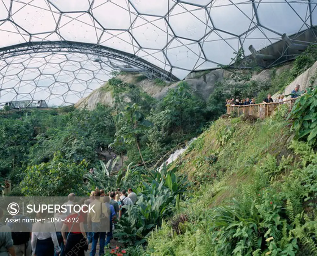 England, Cornwall, St Austell, Eden Project. Tropical Dome Interior With Visitors On Path Leading Past Waterfall