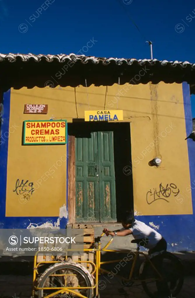 Mexico, Chiapas, San Cristobal De Las Casas, Man Pushing Bicycle Handcart Stacked With Crates Of Fruit Past Yellow And Blue Painted Building Facade With Advertisment For Shampoo Beside Green Door.