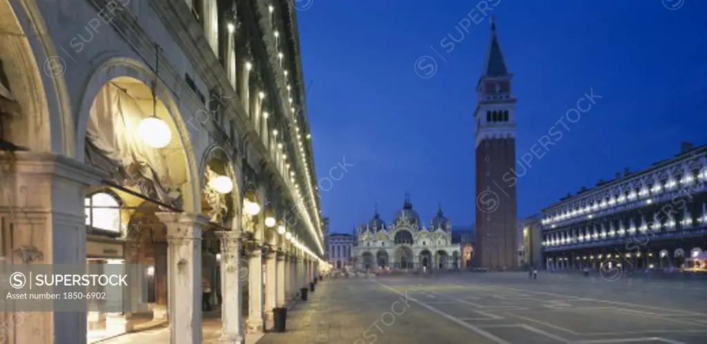 Italy, Veneto, Venice, Piazza San Marco.  View Along The Colonnaded Procuratie Vecchie Towards The Basilica Di San Marco And The Campanile Illuminated At Dusk.