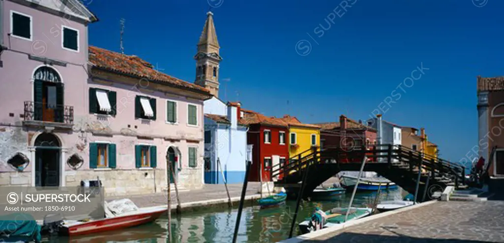 Italy, Veneto, Venice, 'Burano Island.  Bridge And Canal With Boats Moored To Posts Along Each Side Overlooked By Pink, Blue, Red And Yellow Buildings And Bell Tower Behind.'