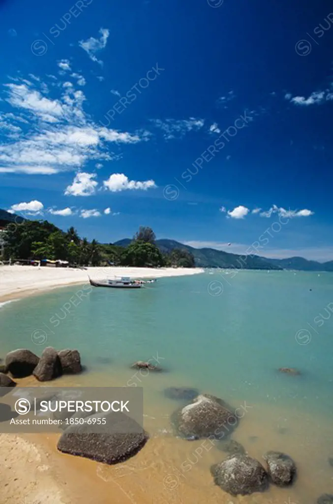 Malaysia, Penang, Batu Ferringhi, View Along Shore Of Quiet Sandy Beach With Moored Boat In Middle Distance And Smooth Rocks In Shallow Water In The Foreground.