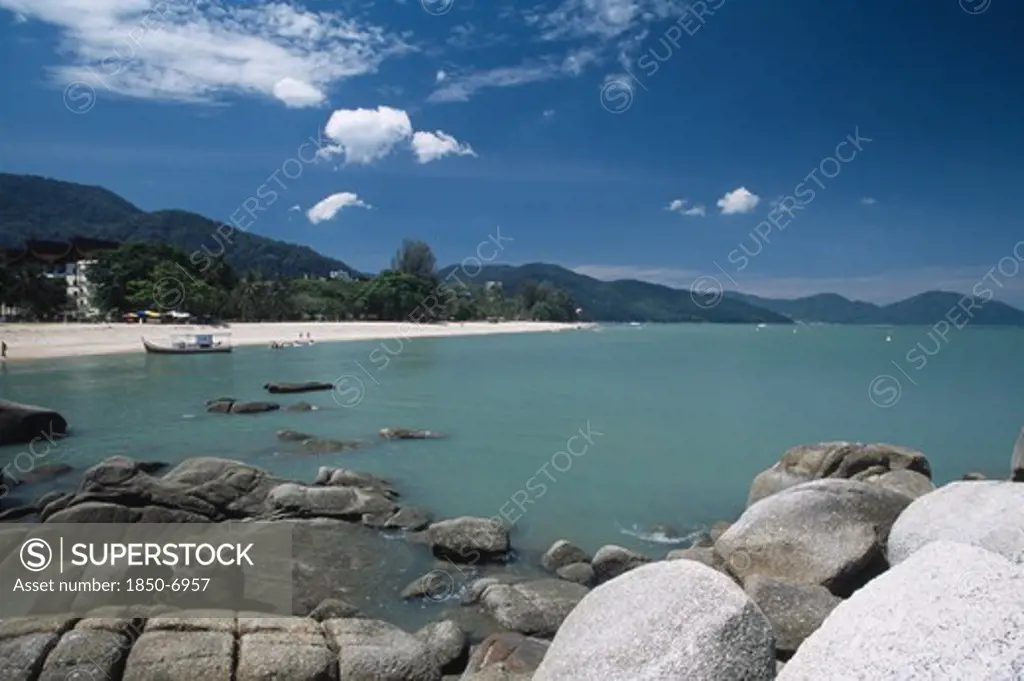 Malaysia, Penang, Batu Ferringhi, View Over Rocks And Bay Of Turquoise Water Towards Distant Moored Boat And Quiet Sandy Beach.