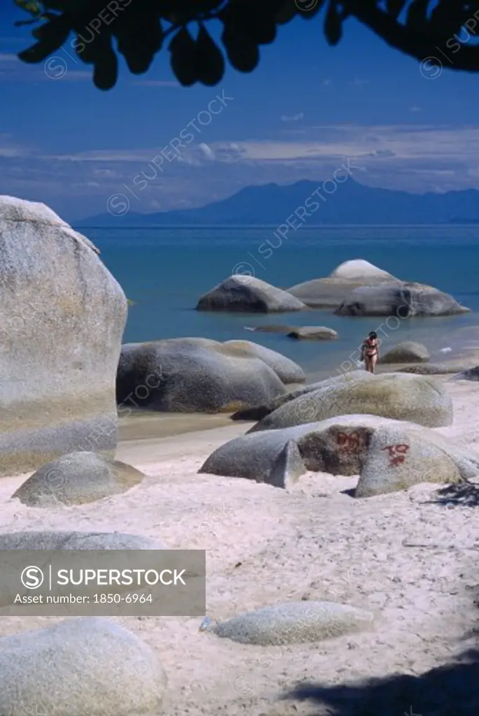 Malaysia, Penang, Batu Ferringhi, 'Sandy Beach With Woman In Bikini Walking Out Of Sea Between Large, Smooth Rocks, Partly Framed By Silhouetted Tree Branch'