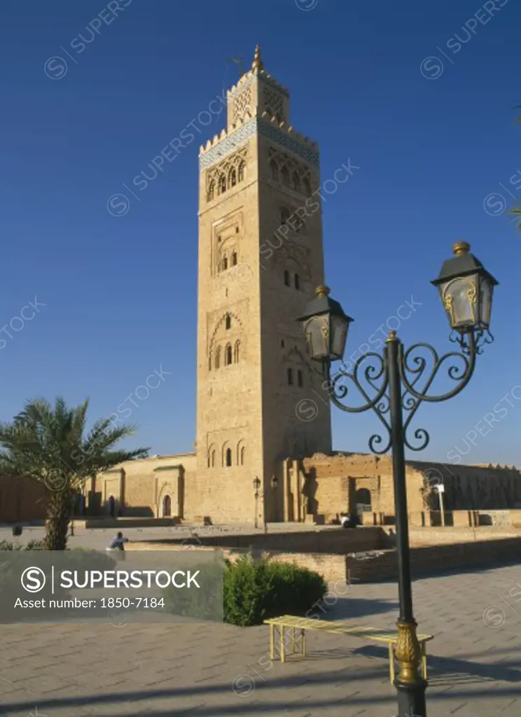 Morocco, Marrakech, Koutoubia Mosque Tower Seen From Pavement With Blue Sky Behind And Street Lamps In Foreground