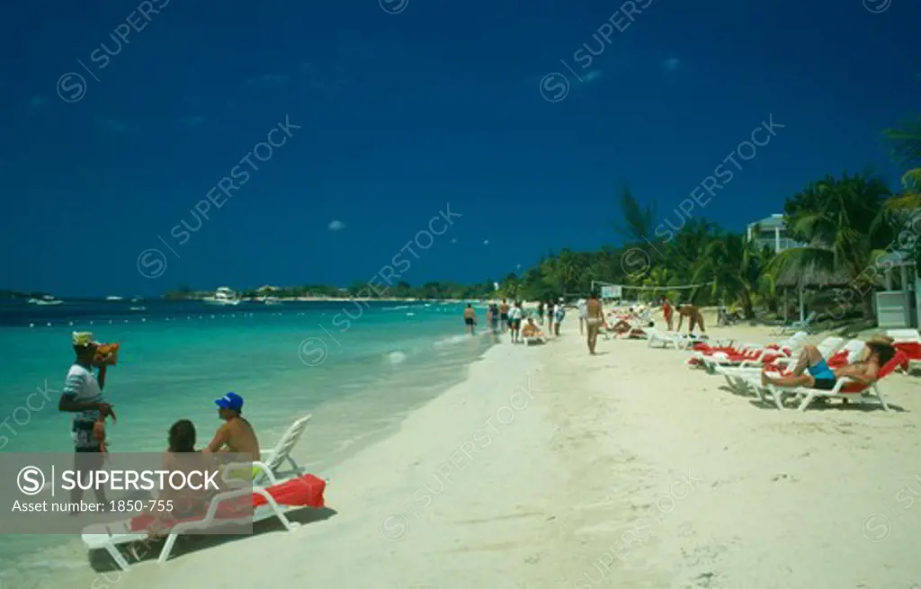 West Indies, Jamaica, Negril, Man Selling Lobster To Tourists On Beach Sitting On Sun Loungers By The Water With Other Tourist Sunbathing