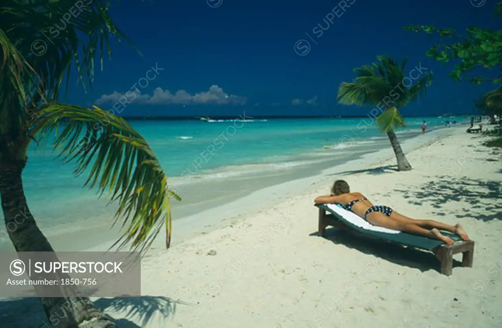 West Indies, Jamaica, Negril, Woman Sunbathing On Lounger On Beach Near Water And Coconut Palm Trees