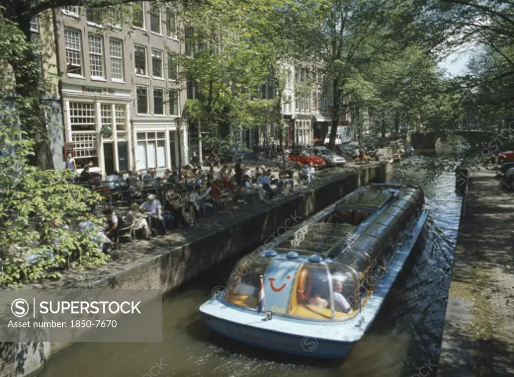 Holland, Noord, Amsterdam, Glass Roofed Tourist Boat Passing Canal Side Cafe On The Lelegracht Canal