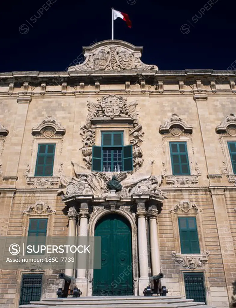 Malta, Valletta, Auberge De Castille Et Leon. Decorative Baroque Facade With Steps Leading To Green Doorway Flanked By Pillars And Cannons With The Maltese Flag Flying From Roof. Official Residence Of The Prime Minister