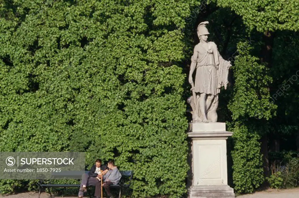 Austria, Vienna, Schonbrunn Gardens.  Classical Statue Against Hedge With Two Visitors Sitting On Bench At One Side.