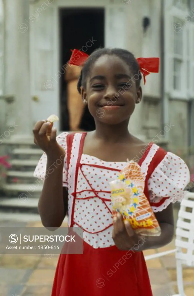 West Indies, Barbados, Easy Hall, Young Smiling Girl In Red And White Dress With Red Ribbons In Hair Eating Crisps From A Packet