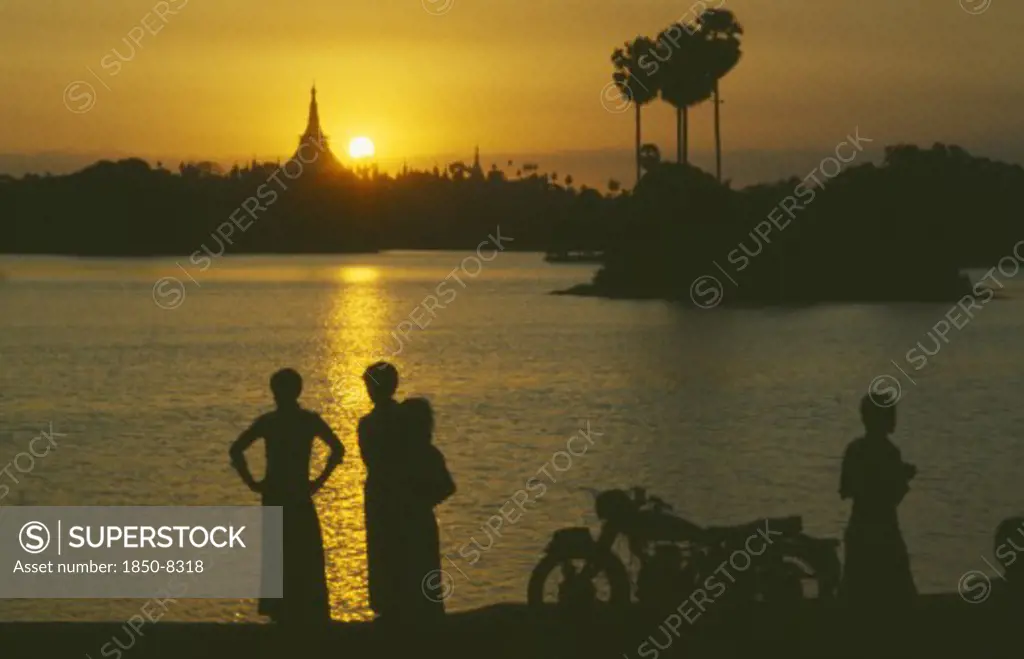 Myanmar, Yangon, People Silhouetted On Shore Of Lake Looking Across Towards The Shwedagon Pagoda At Sunset.