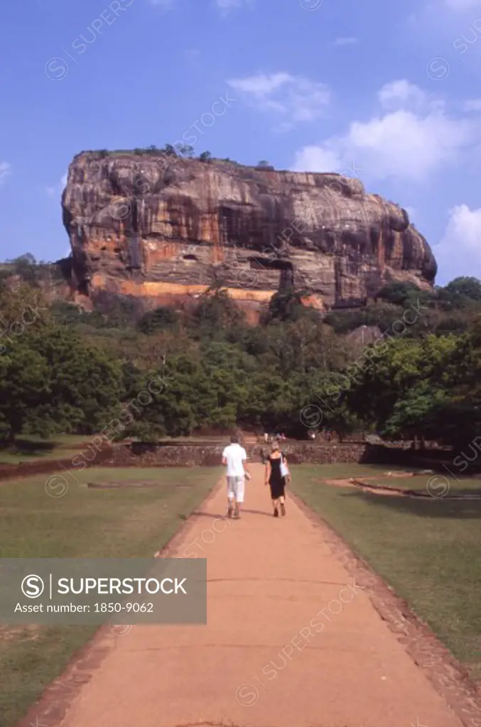 Sri Lanka, Sigiriya, View Towards Huge Monolithic Rock Site Of Fith Century Citadel.  Also Called Lion Rock.  Tourist Couple On Path Through Gardens In The Foreground.