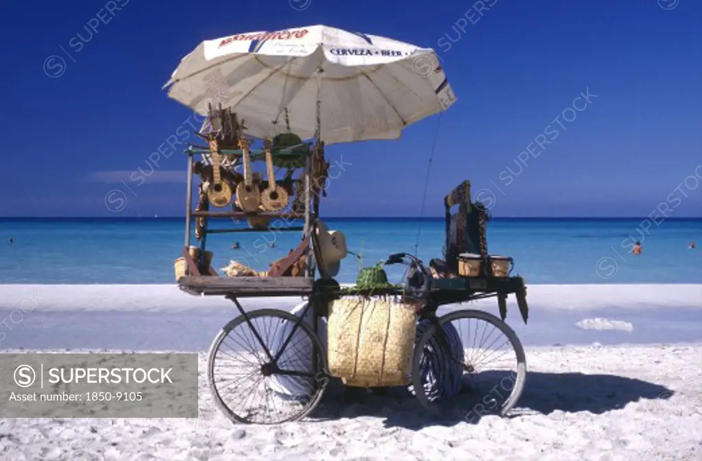 Cuba, Varadero, Beach Vendors Stall On A Bycicle Selling Musical Instruments And Hats With Clear Blue Sea Beyond