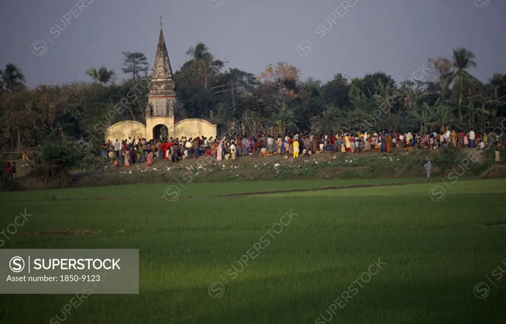Bangladesh, Aricha, View Over Rice Fields Towards Crowds Gathered Around Old Hindu Temple At Sunset.