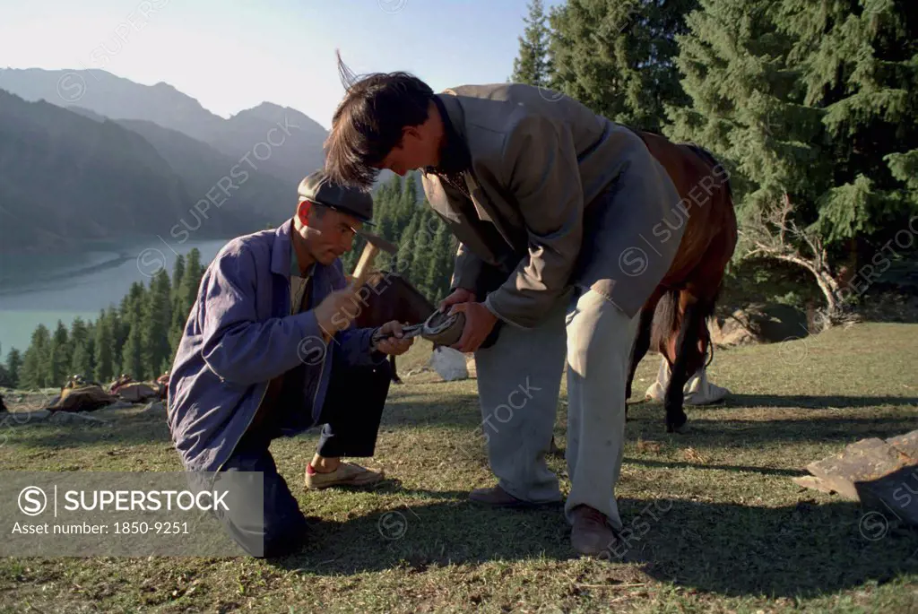 China, Xinjiang, Tianchi, Heavenly Lake. Two Men Shoeing A Horse Near The Lakeside