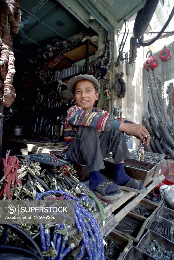 China, Xinjiang, Kashgar, Young Hardware Vendor Sitting Amongst Goods For Sale
