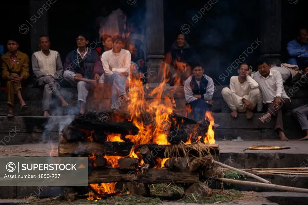 Nepal, Kathmandu, Hindu Cremation In Pashupatinath Temple.  Burning Wood Funeral Pyre With Onlookers On Steps Behind.