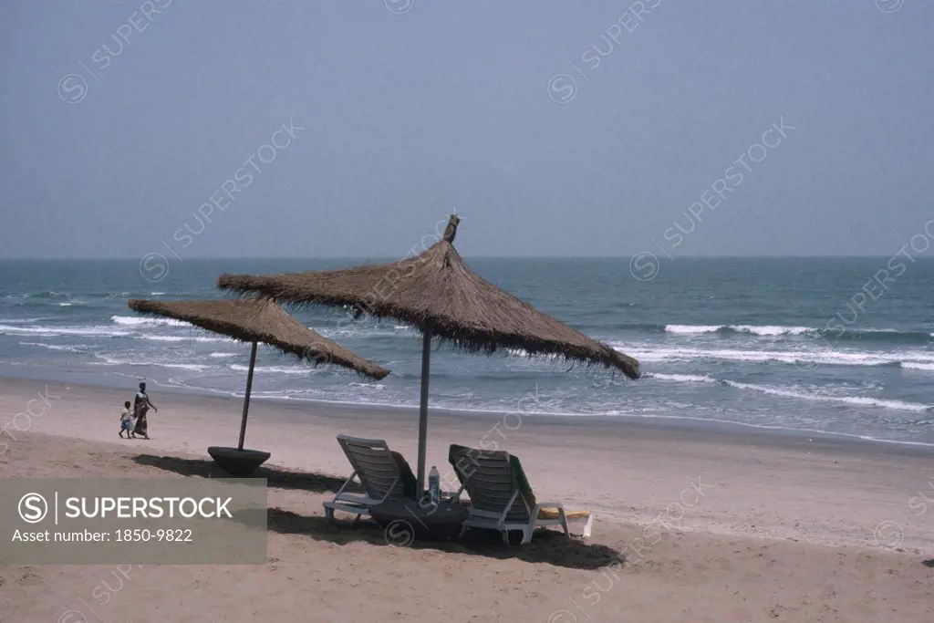 Gambia, People, Hotel Beach Front With Woman And Child Walking Hand In Hand Over Sand. Sun Loungers And Parasols In The Foreground