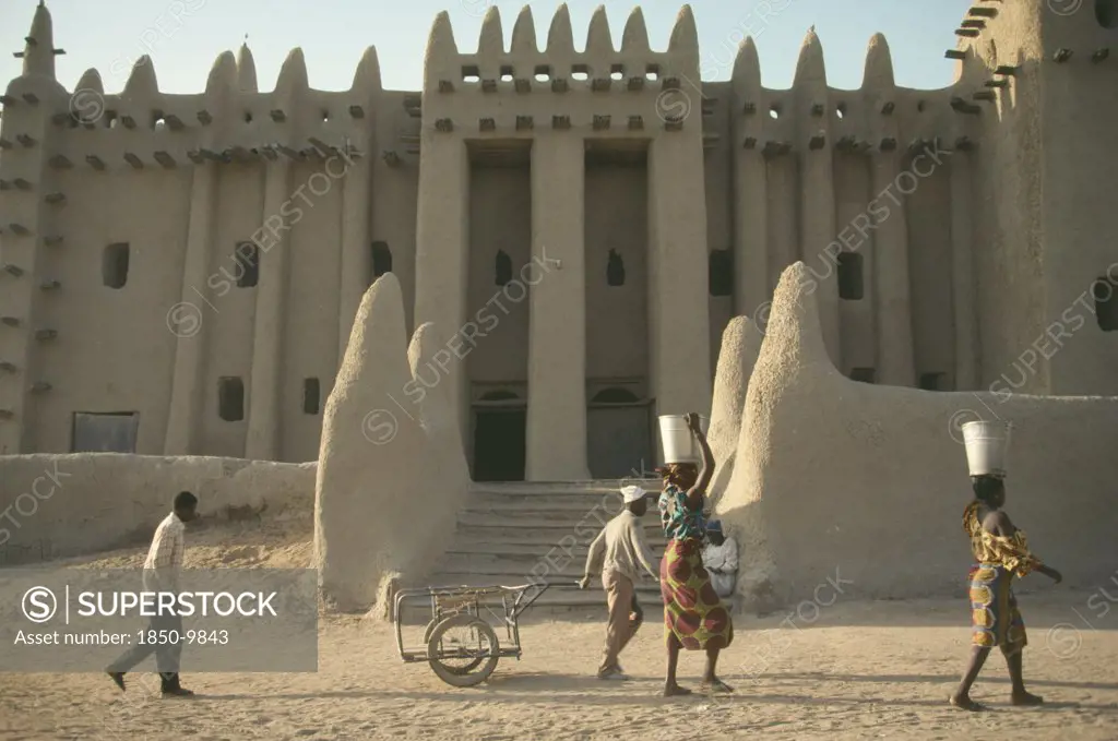 Mali, Djenne, Exterior Of Mud Brick Mosque Built In Traditional Style With Woman Walking Past Carrying Buckets On Their Heads.