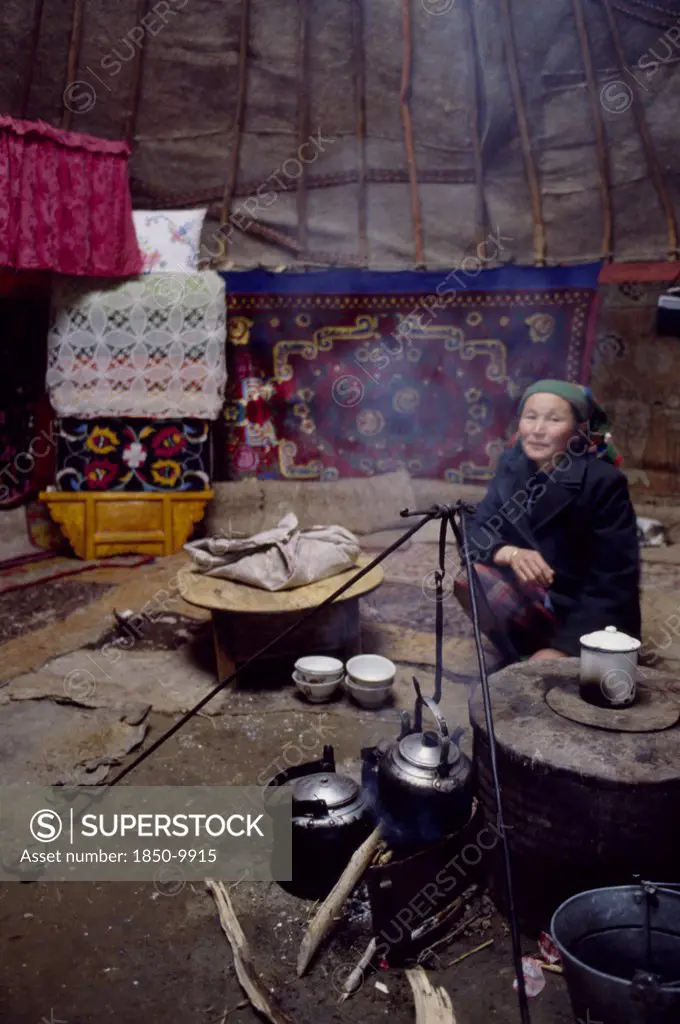 China, Xinjiang Province, Altai Region, Kazakh Woman Inside Her Kigizuy Or Yurt With Covered Stove And Kettle Suspended Over Small Open Wood Fire.