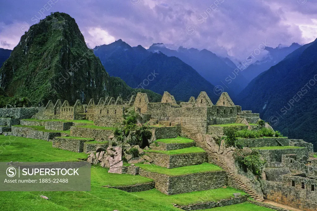 Peru, Inca ruins at Maccu Piccu - showing central courtyard and Huayna Piccu landmark, grass terraces, ancient stonework - dark clouds and mountain valley peaks in background