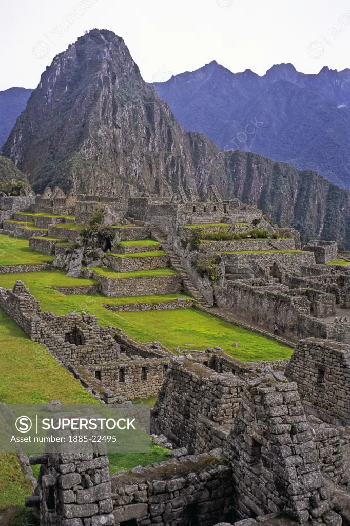 Peru, Inca ruins at Maccu Piccu - showing central courtyard and Huayna Piccu landmark, grass terraces, ancient stonework - dark clouds and mountain peaks in background, tourist in view