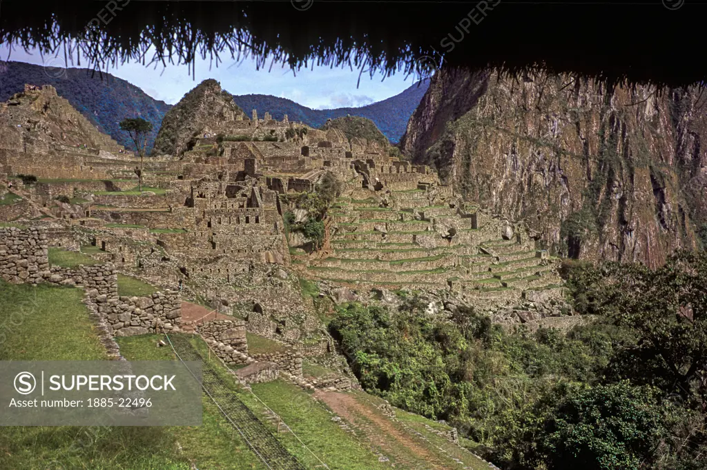 Peru, Inca ruins at Maccu Piccu - showing central courtyard and Huayna Piccu landmark, grass terraces, ancient stonework - dark clouds and mountain peaks in background, tourist in view, with thatched roof on top edge