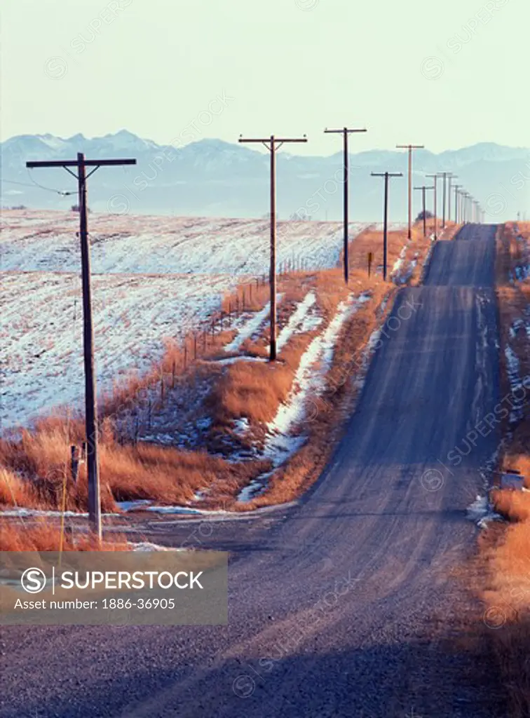 Gravel road lined by telephone or power poles through the farmland and snowy wheat fields, winter in Gallatin Valley, Montana, USA.